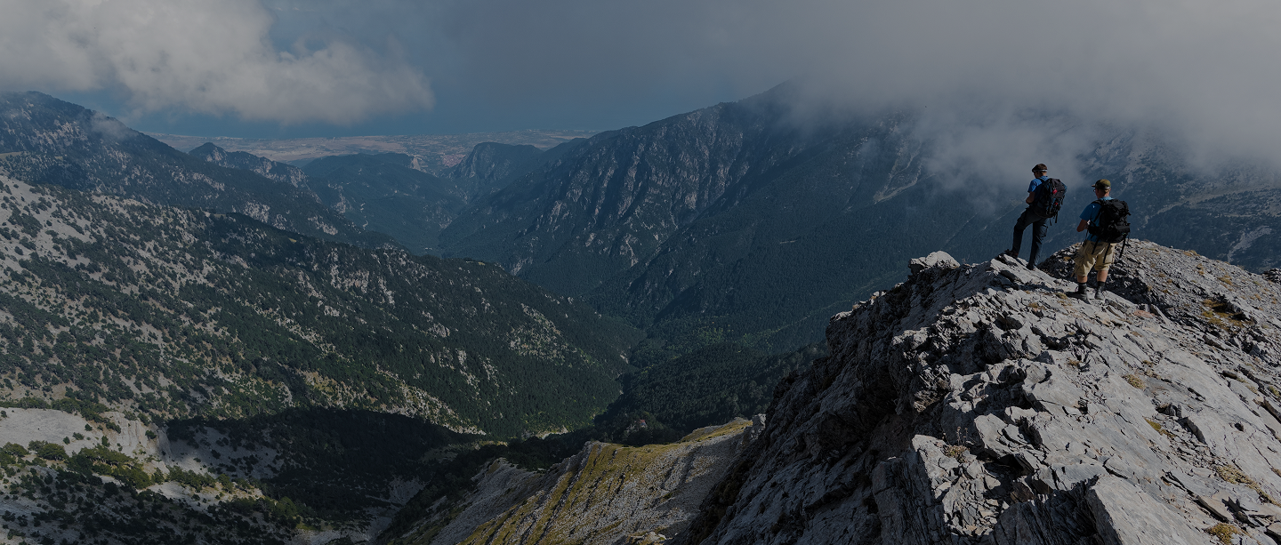 Two hikers with full packs standing on a mountain peak looking into valley below surrounded by mountains, Banner for the home page